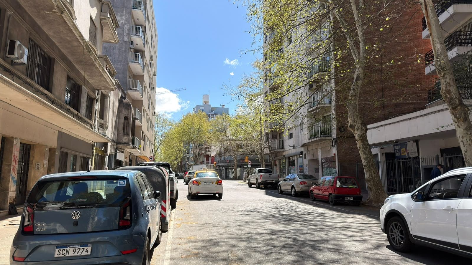 # Street View - Urban Neighborhood
This image shows a charming urban street in what appears to be a South American city (likely Uruguay based on visible license plates), lined with mid-rise residential and mixed-use buildings featuring a blend of architectural styles. The tree-lined pedestrian-friendly street includes parked vehicles, ground-floor commercial spaces, and well-maintained buildings with balconies, offering a vibrant neighborhood atmosphere with excellent natural light and clear blue skies.