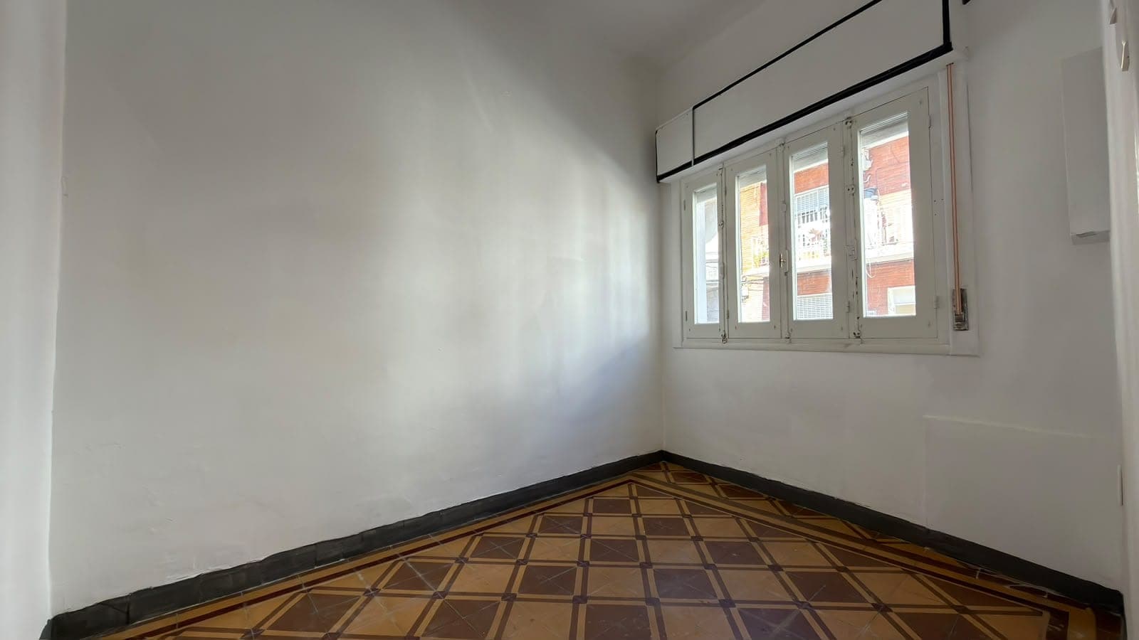 This is a bright, unfurnished bedroom or living space with clean white walls and excellent natural lighting from a large multi-pane window with white frames overlooking neighboring buildings. The room features original geometric patterned tile flooring in warm brown tones with a dark border trim, and a modern black ceiling detail. The space is in good condition and appears ready for occupancy or renovation.